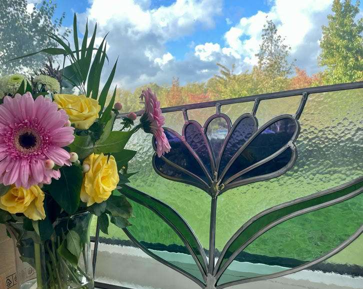 A vase with pink and yellow flowers on a windowsill, next to a stained glass window with a floral design, with a view of a grassy area and trees under a blue sky with clouds.
