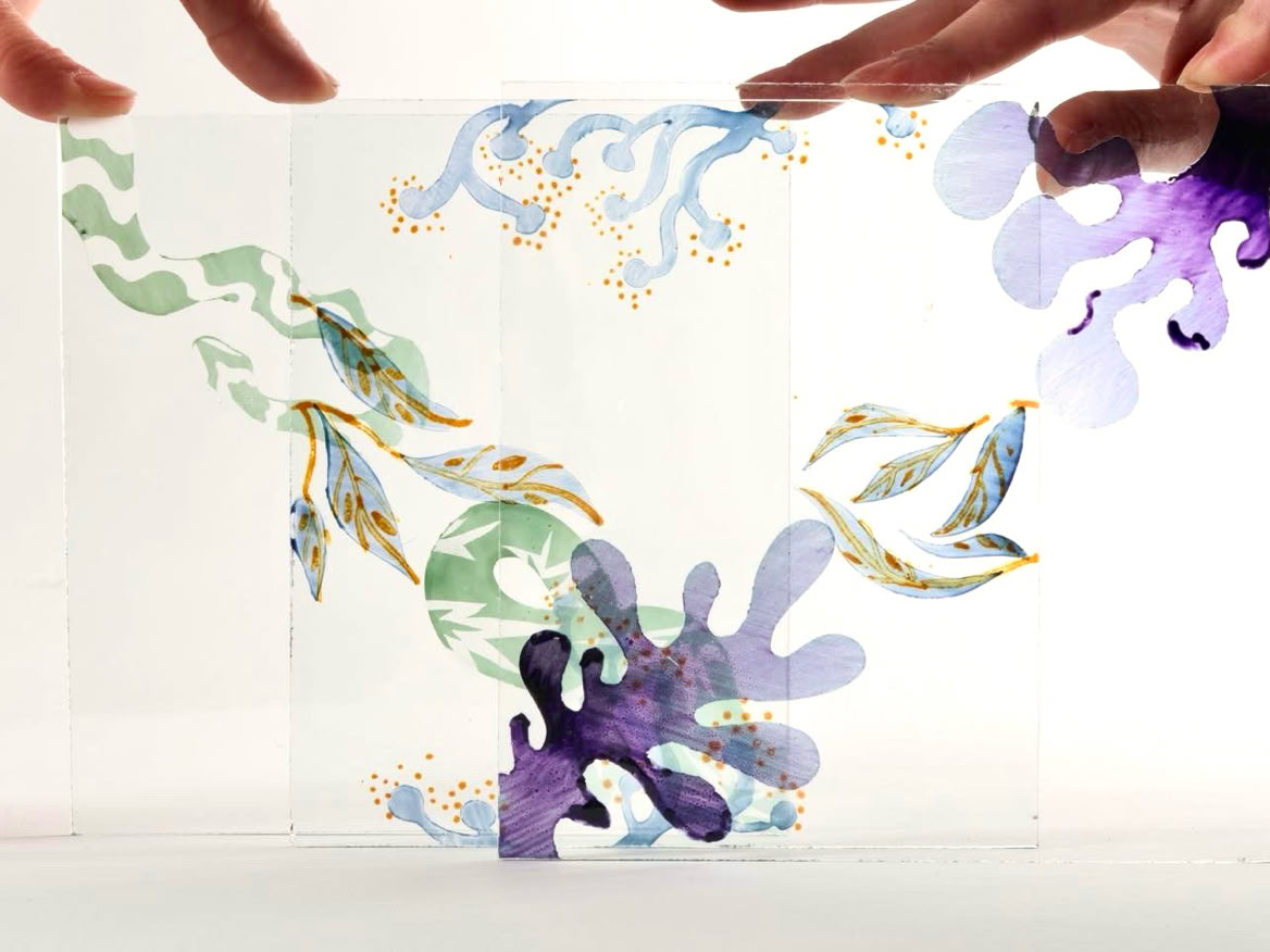 Two hands holding a transparent sheet with colorful abstract designs, including purple, green, and blue organic shapes.