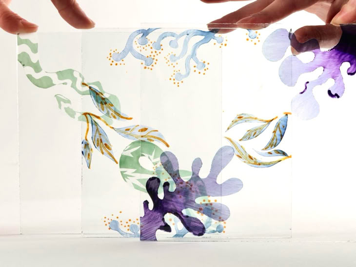 Two hands holding a transparent sheet with colorful abstract designs, including purple, green, and blue organic shapes.