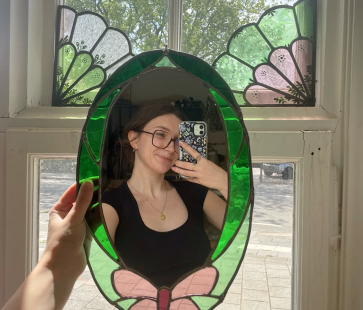 A person holding an oval mirror with a green and pink butterfly design, reflecting their image. The background shows a window with stained glass panels and a view of trees and the sky outside.