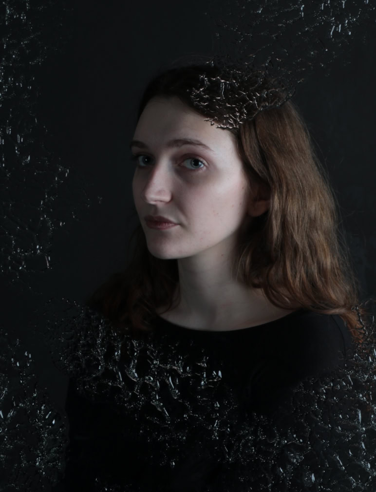 A person with long hair wearing a dark top, standing against a dark background with a textured overlay.