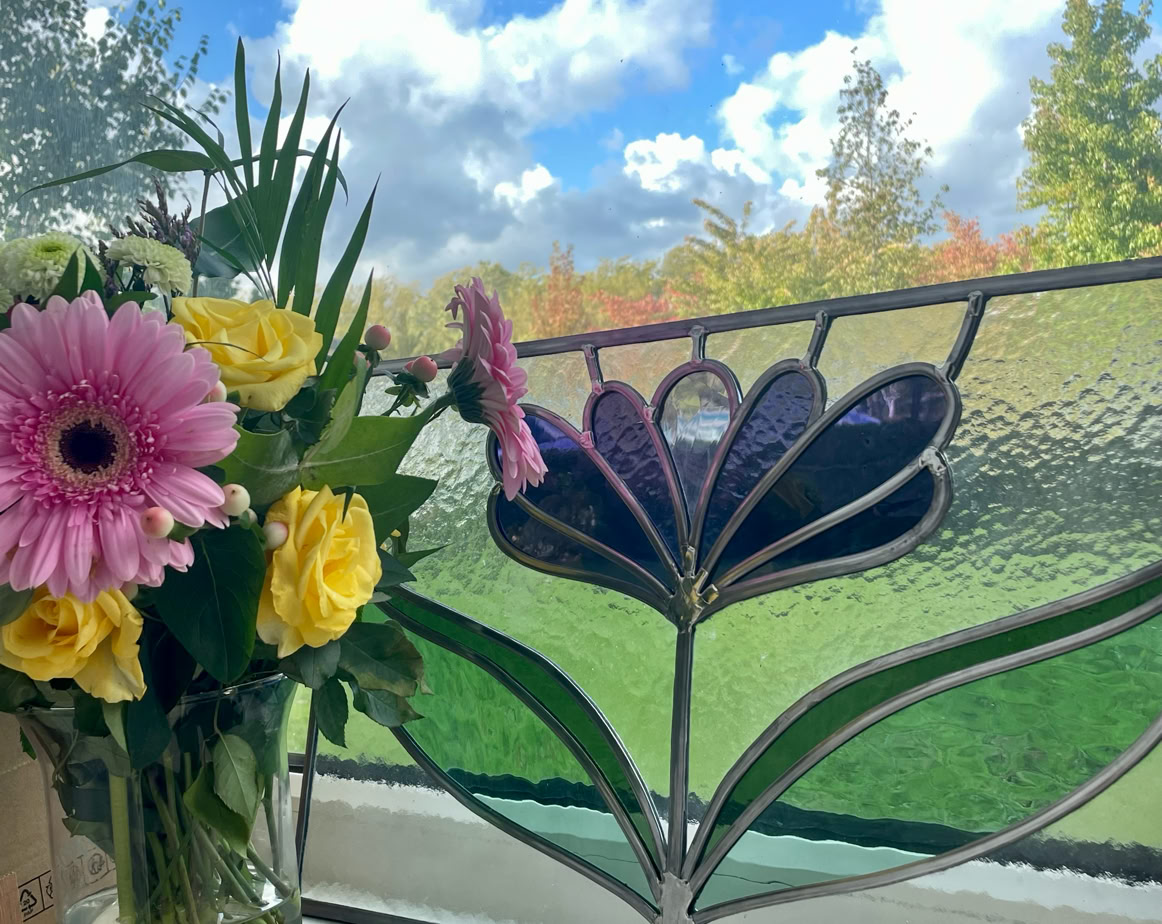 A vase with pink and yellow flowers on a windowsill, next to a stained glass window with a floral design, with a view of a grassy area and trees under a blue sky with clouds.